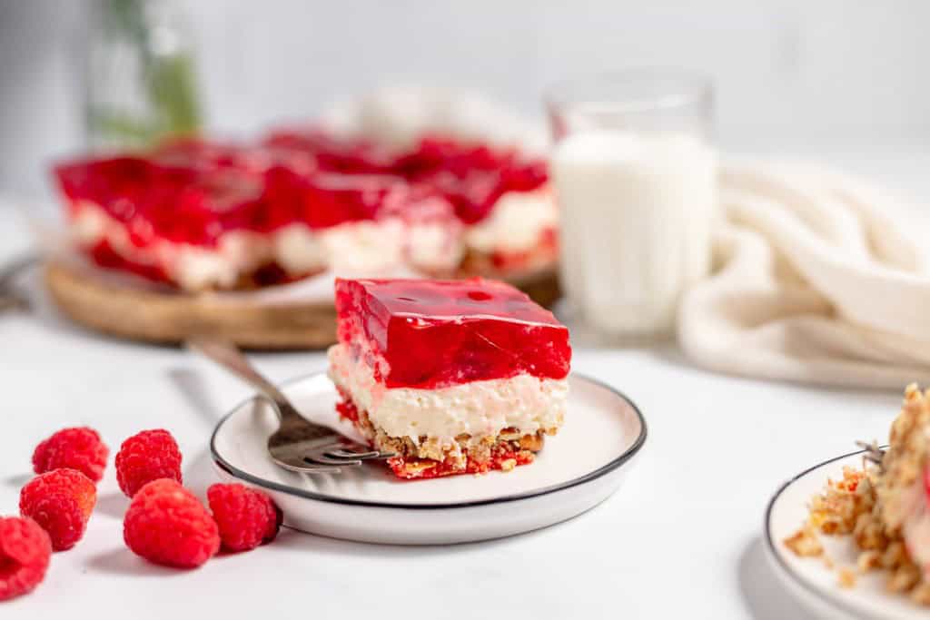A slice of dessert with red gelatin and cream layers on a plate with a fork, surrounded by fresh raspberries, a glass of milk, and a white cloth on a marble surface.