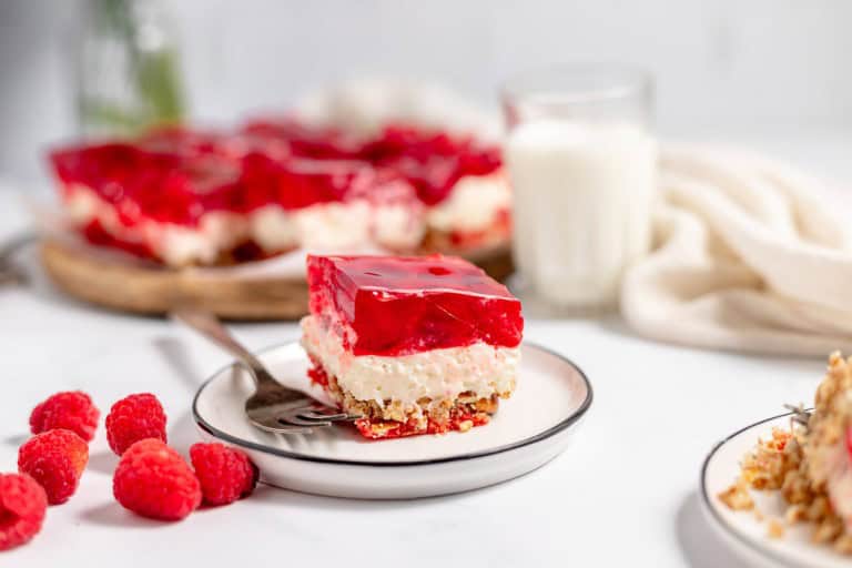 A slice of dessert with red gelatin and cream layers on a plate with a fork, surrounded by fresh raspberries, a glass of milk, and a white cloth on a marble surface.