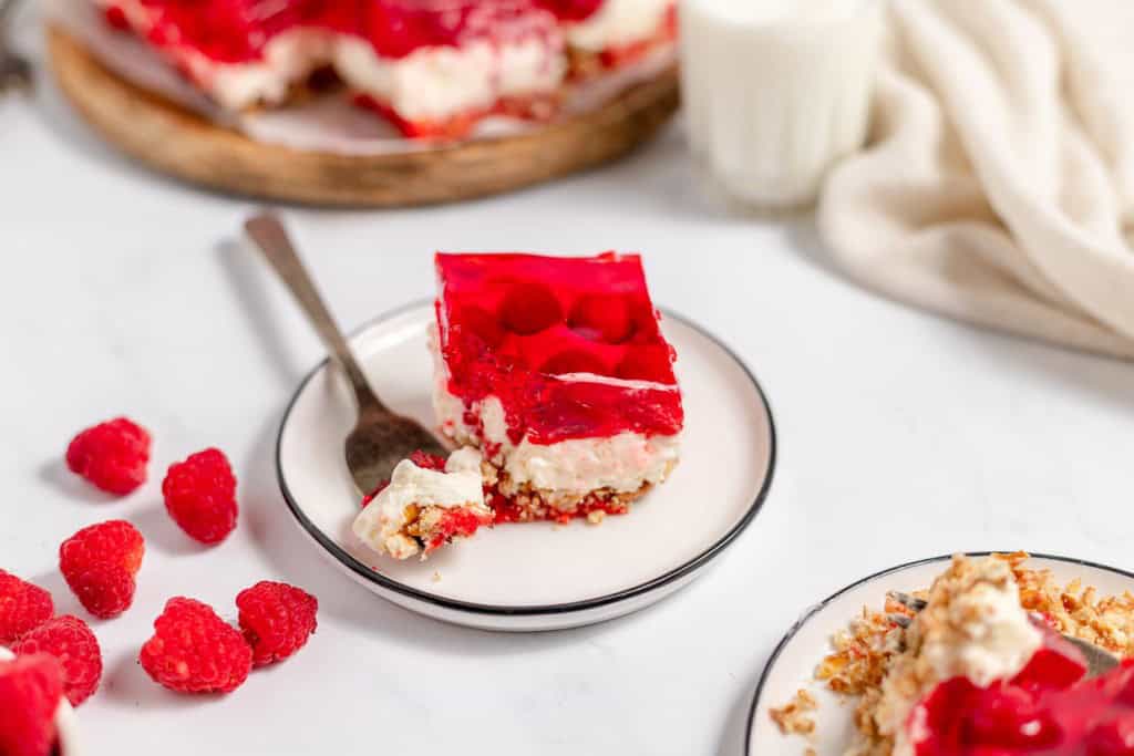 A slice of raspberry pretzel salad with a fork on a plate, surrounded by fresh raspberries and garnished with a napkin, with another plate of dessert and a glass of milk in the background.