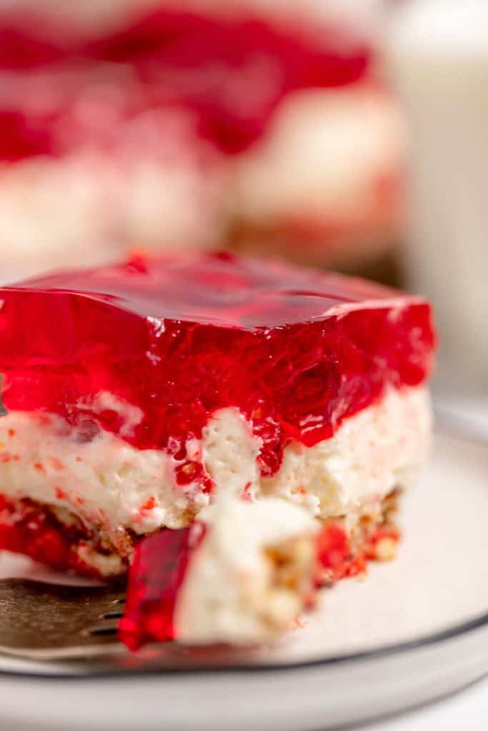 Close-up of a dessert bar with layers of red gelatin, creamy filling, and a crumbly base, showing a bite taken out with a fork.