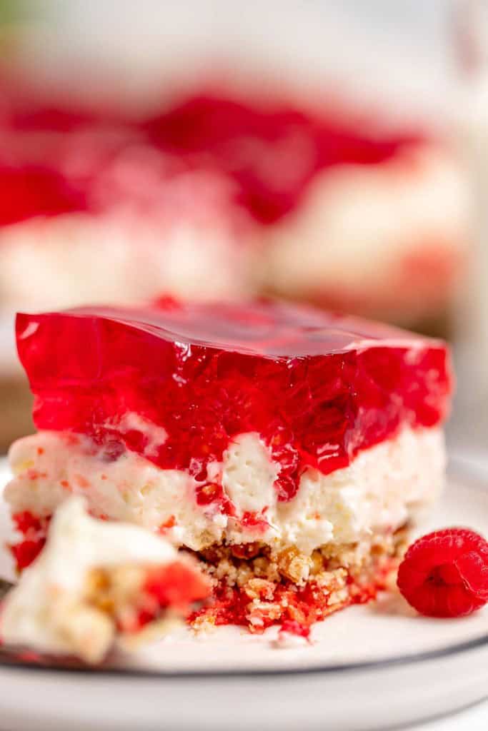 A slice of layered dessert comprising a crumb crust, creamy middle layer, and red gelatin top, garnished with a raspberry, placed on a white plate.