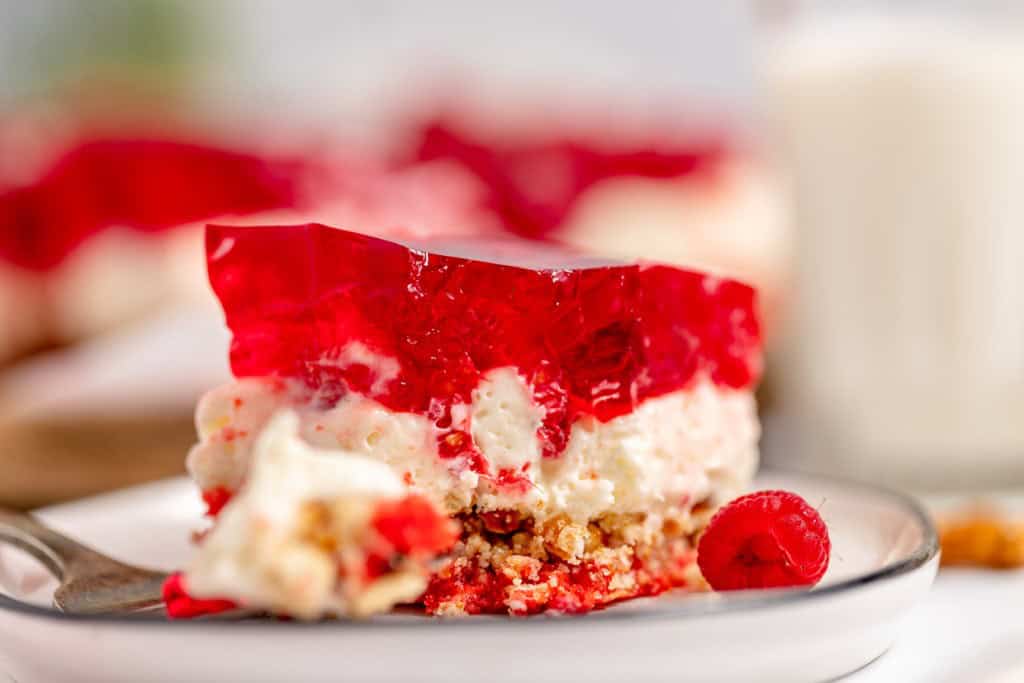 A dessert slice with layers of red gelatin, creamy filling, and a crumbly crust, served on a white plate with a raspberry on the side.