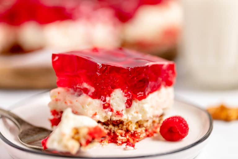 A plate with a slice of strawberry pretzel salad, featuring a pretzel crust, creamy middle layer, and strawberry gelatin topping, with a fork and a raspberry on the side.