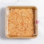 A square baking pan filled with layered crumb mixture lined with parchment paper and secured with a pink binder clip on one side, placed on a marble surface.