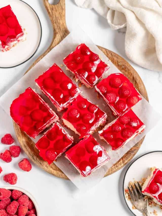 Top-down view of raspberry-topped dessert bars on a wooden serving tray, surrounded by a bowl of raspberries and a white cloth.