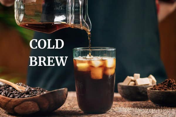 A person pours cold brew coffee from a pitcher into a glass with ice. Nearby are coffee beans, sugar cubes, and ground coffee. The words "COLD BREW" are displayed beside the glass.