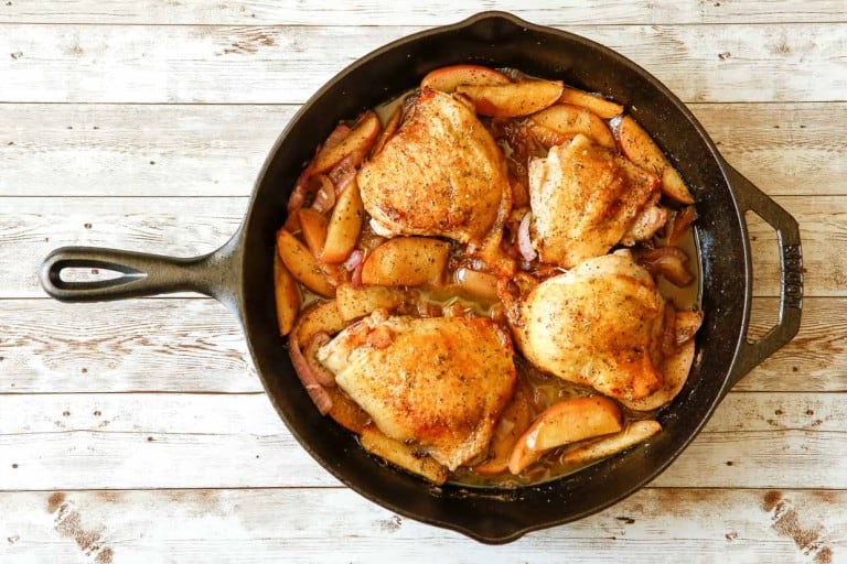 A cast iron skillet dinner featuring four pieces of browned chicken thighs paired with sliced apples, all set on a rustic wooden surface.