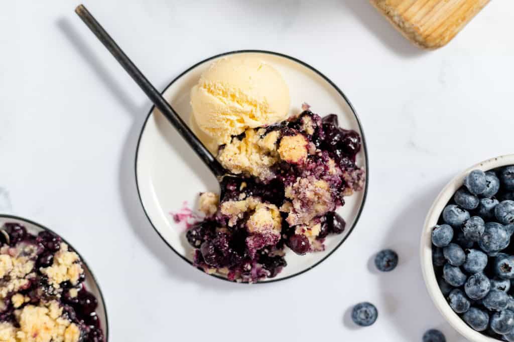 A dessert plate with vanilla ice cream and blueberry crisp topped with a spoon, surrounded by fresh blueberries.