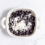 A white ceramic dish filled with blueberries and a dusting of white flour on top, set on a marble surface.