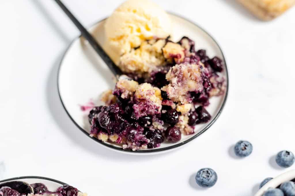 A dessert plate with a serving of blueberry crumble topped with a scoop of vanilla ice cream, accompanied by a spoon. Blueberries are scattered around the plate on a white surface.