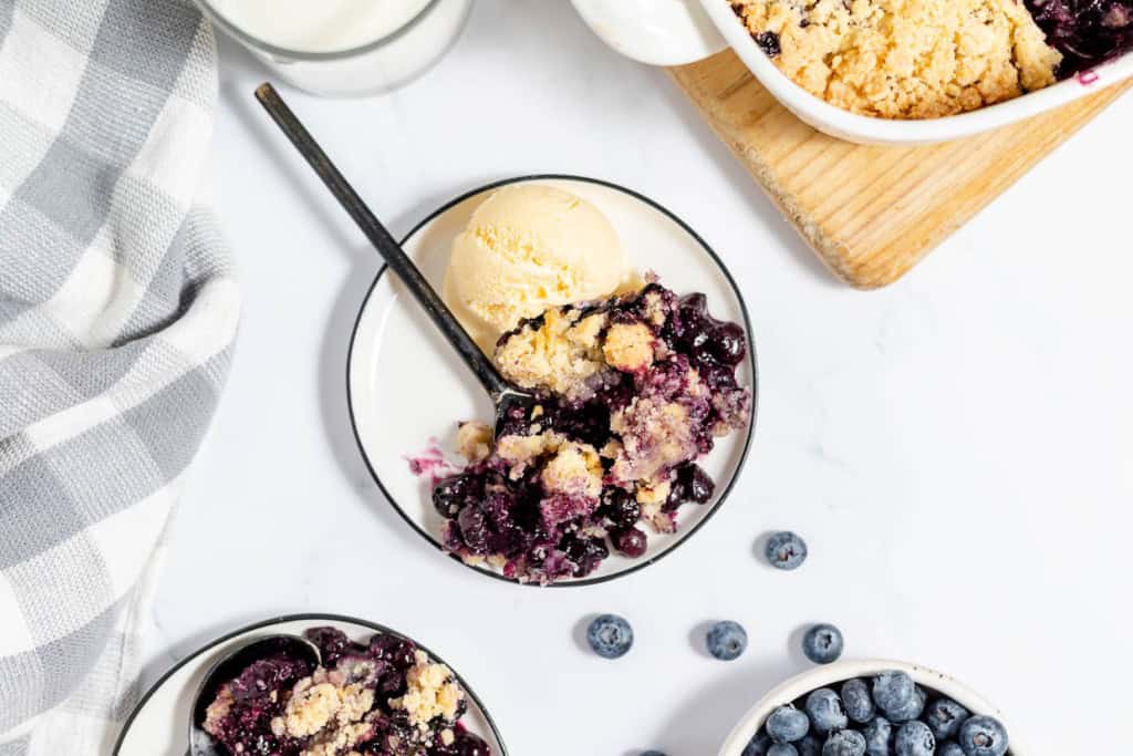 A plate with a serving of blueberry crumble and a scoop of vanilla ice cream, with blueberries, a checked cloth, and a bowl of crumble in the background.