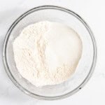 A clear glass bowl containing white powder, possibly flour or a similar baking ingredient, on a white marble surface.