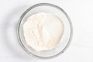 A clear glass bowl containing white powder, possibly flour or a similar baking ingredient, on a white marble surface.