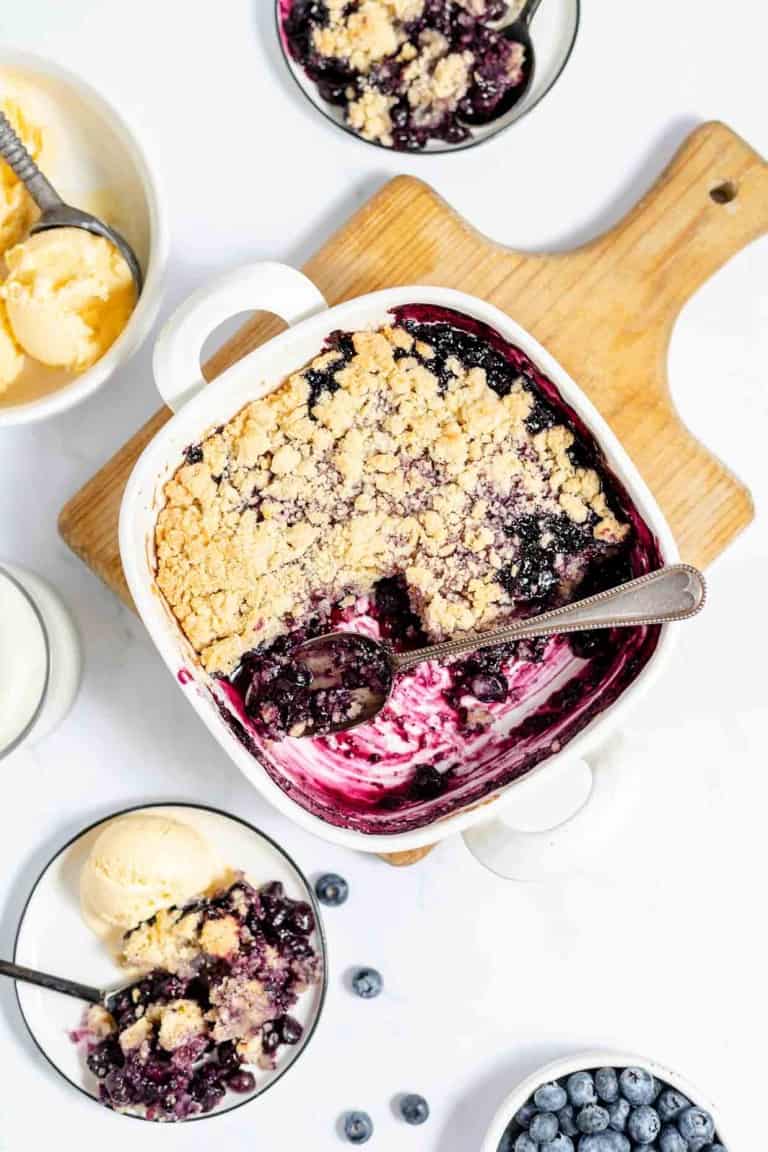 A partially eaten blueberry crumble in a white dish with a spoon, surrounded by bowls of vanilla ice cream, fresh blueberries, and a glass of milk on a wooden board.