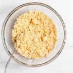 A glass bowl filled with crumbly mixture and a spoon placed on a white surface.