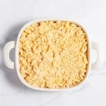 A white rectangular baking dish filled with uncooked, crumbly yellow topping sits on a white marble surface.