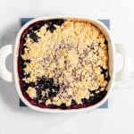 A white baking dish filled with blackberry crumble, featuring a golden-brown crumbly top layer and dark purple-blackberry filling beneath. The dish sits on a blue coaster on a white surface.