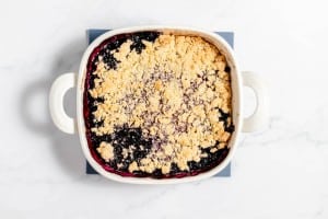 A white baking dish filled with blackberry crumble, featuring a golden-brown crumbly top layer and dark purple-blackberry filling beneath. The dish sits on a blue coaster on a white surface.