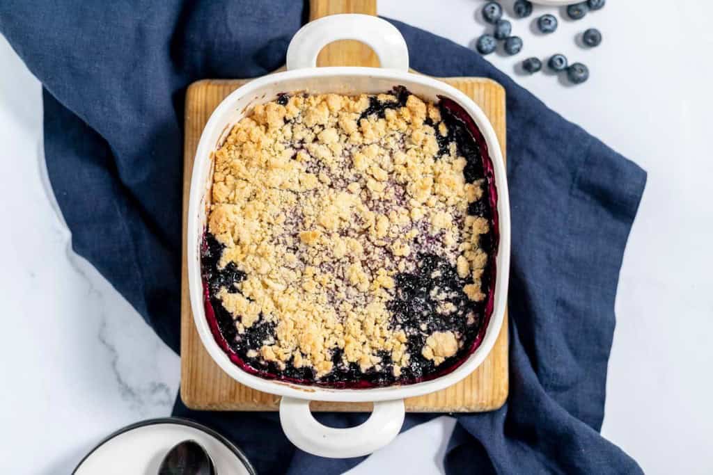 A square baking dish filled with blueberry crisp topped with a crumbly golden brown streusel, placed on a wooden board with a blue cloth and scattered blueberries beside it.