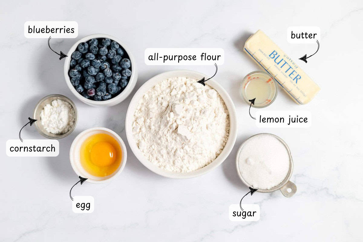 Ingredients for a delightful Blueberry Crumble on a white surface: fresh blueberries, all-purpose flour, butter, cornstarch, egg, lemon juice, and sugar.