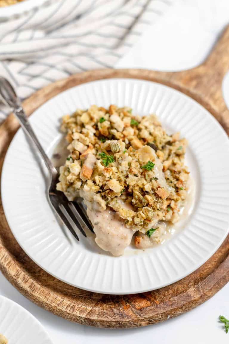 A rectangular white baking dish filled with a delicious Chicken and Stuffing Casserole topped with breadcrumbs. A serving spoon rests on the left side, with a portion of the casserole missing. A striped cloth lies next to the dish.