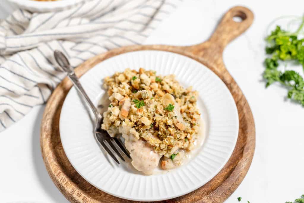 A white plate with a wooden base holds a serving of delicious Chicken and Stuffing Casserole, garnished with fresh parsley, beside a fork. A striped cloth and parsley garnish are visible in the background.