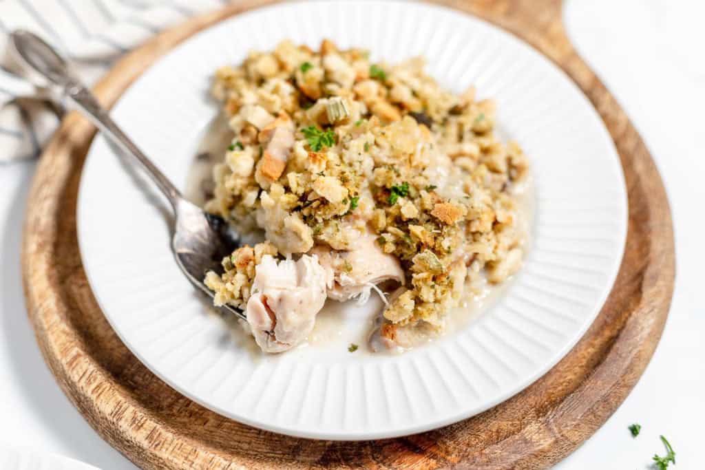 A white plate with a serving of Chicken and Stuffing Casserole, garnished with herbs, placed on a wooden board. A fork is resting on the plate.