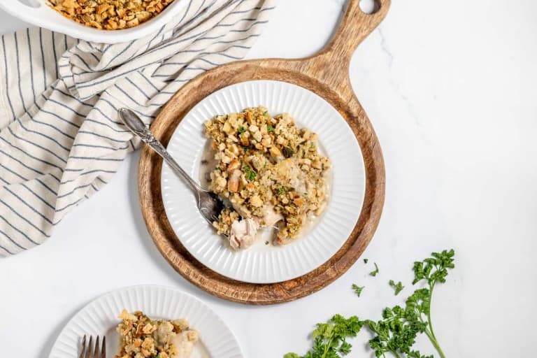 A plated meal of chicken and stuffing casserole is placed on a wooden board next to a fork and a striped cloth. Fresh parsley and a partially seen casserole dish are in the background.