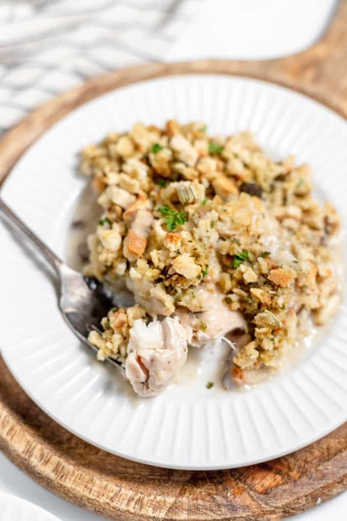 A plate with a serving of Chicken and Stuffing Casserole, garnished with herbs, on a wooden tray. A fork is placed on the plate, partially filled with a bite of the dish.