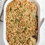 A white casserole dish filled with a savory Chicken and Stuffing Casserole, topped with golden breadcrumbs, with a spoon partially removing a portion. A striped cloth is beside the dish.