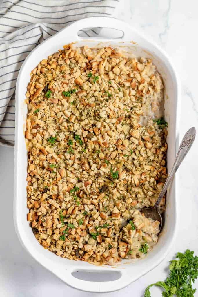 A white casserole dish filled with a savory Chicken and Stuffing Casserole, topped with golden breadcrumbs, with a spoon partially removing a portion. A striped cloth is beside the dish.