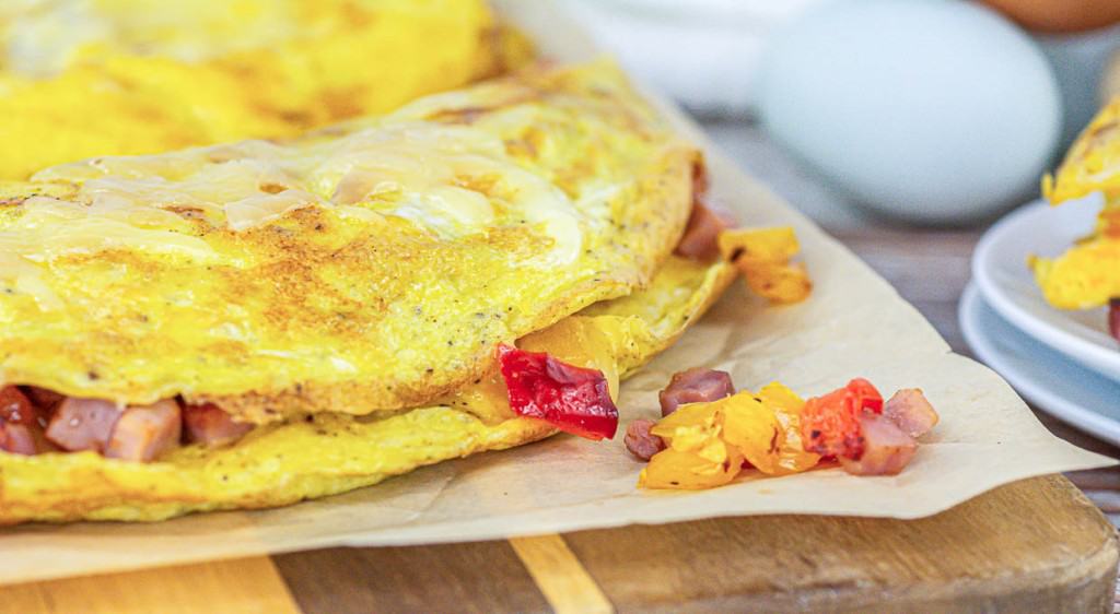 Close-up of an omelet filled with diced ham and bell peppers placed on parchment paper, with eggs and a partially visible plate in the background&mdash;perfect for your breakfast ideas.
