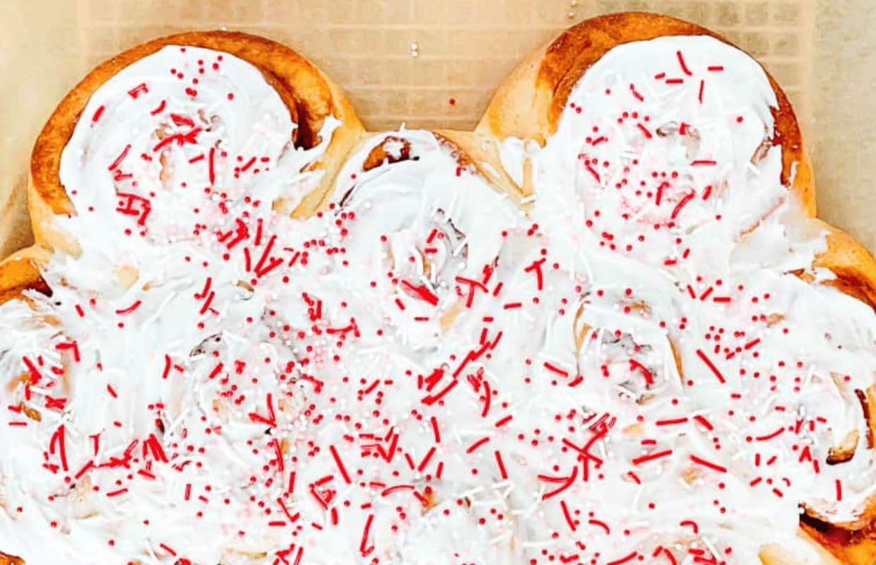 A batch of cinnamon rolls covered in white icing and topped with red sprinkles on a baking sheet.