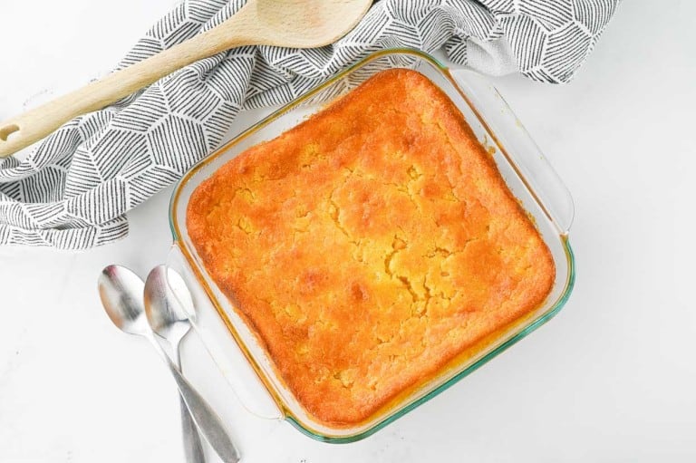 A glass baking dish filled with freshly baked cornbread, a delightful addition to your side dishes, sits next to a patterned cloth, two wooden spoons, and three metal spoons on a white surface.