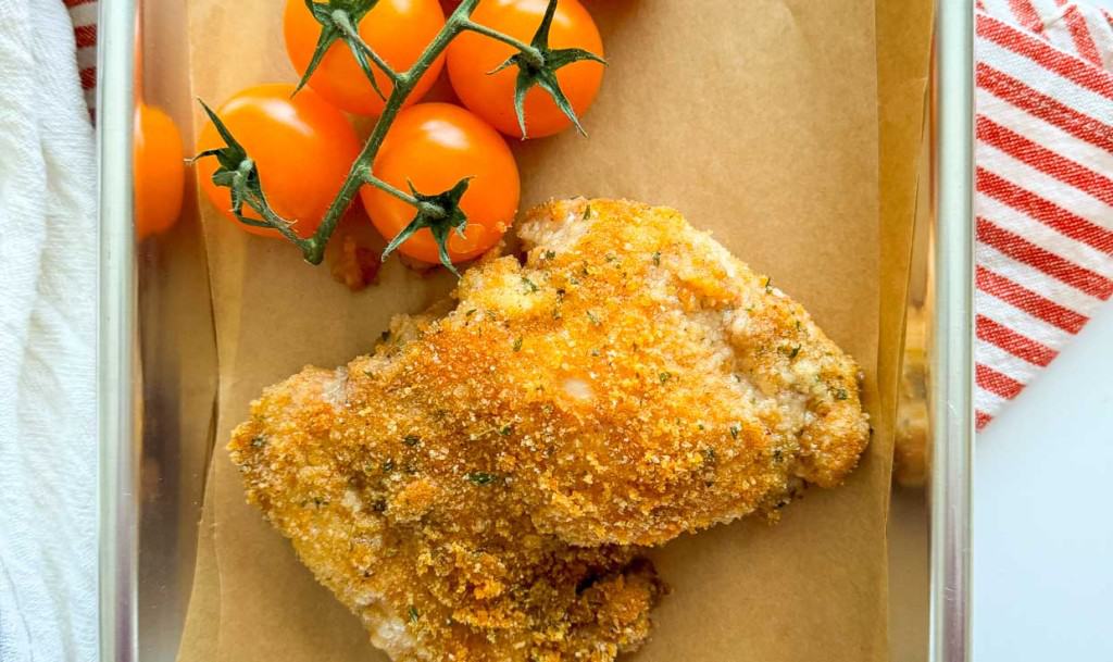 A piece of fried chicken with a twist, breaded to perfection, rests on brown parchment paper next to a vine of ripe orange tomatoes in a clear dish.