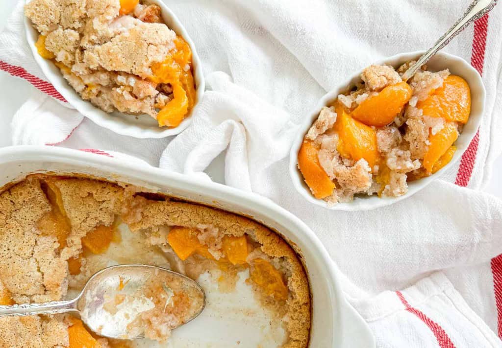 A baking dish with peach cobbler and a spoon, alongside two smaller servings of the cobbler on white linens with red stripes—a delightful addition to your stone fruit recipes.