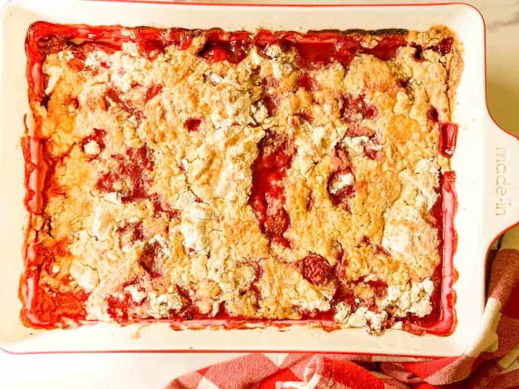 A rectangular baking dish filled with a baked dessert resembling a fruit cobbler, with a mix of golden, crumbly topping and visible patches of red fruit filling. A red and white cloth is partially visible.