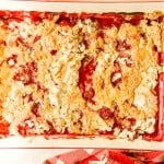 A rectangular baking dish filled with a baked dessert resembling a fruit cobbler, with a mix of golden, crumbly topping and visible patches of red fruit filling. A red and white cloth is partially visible.