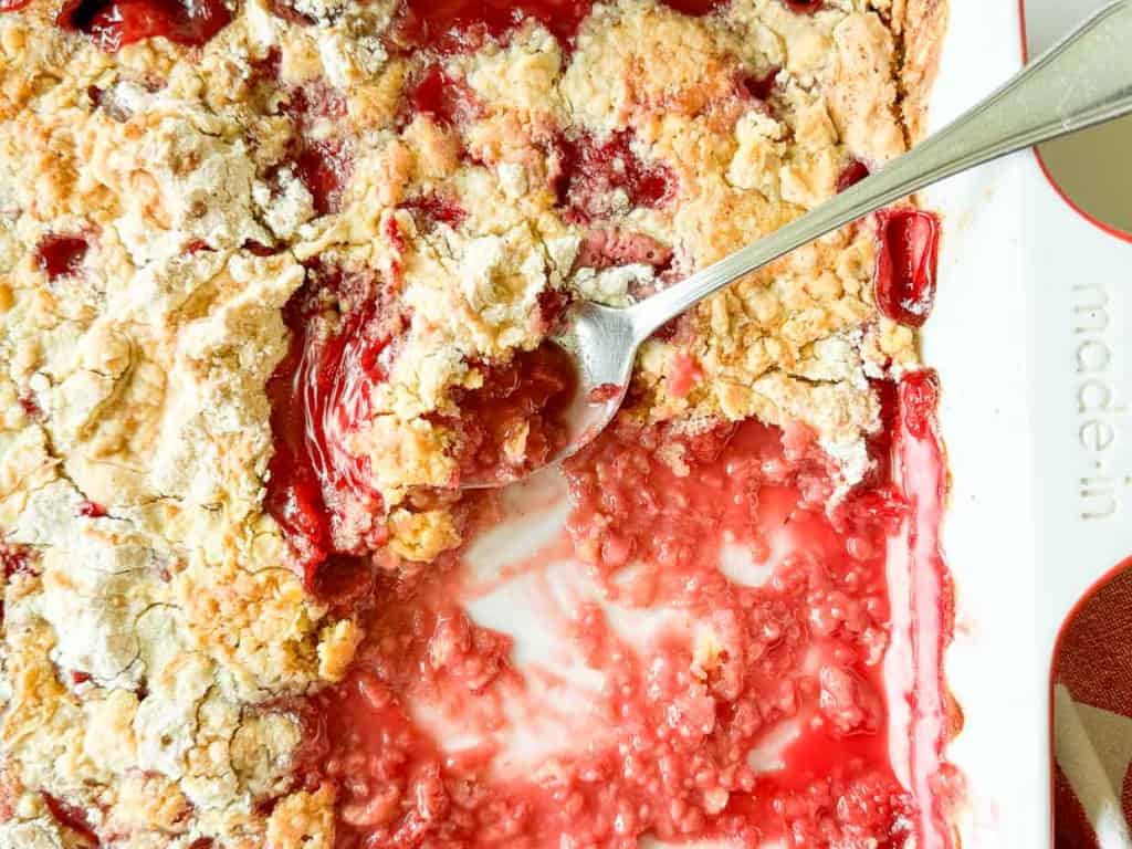 A close-up of a cherry cobbler with a serving spoon scooping out a portion, revealing the red filling and crumbly topping in a white dish.