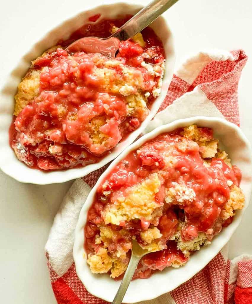 Two white oval dishes filled with a pink and yellow dessert, possibly berry cobbler, each with a spoon. The dishes rest on a red and white checkered cloth.