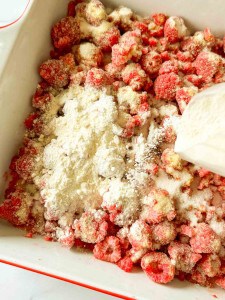 A close-up of a baking dish filled with raspberries covered in white flour and sugar, being mixed together with a white mixing tool.