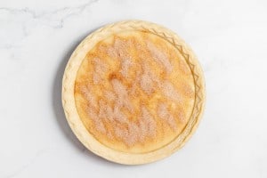 An unbaked pie with a crimped edge crust, filled with a mixture and topped with a sprinkled layer of cinnamon and sugar, on a marbled white surface.