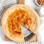 A pie on a wooden board with a slice removed, revealing its interior. A striped cloth, cinnamon sticks in a bowl, and a glass of milk are placed nearby.