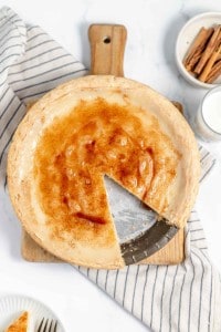 A pie on a wooden board with a slice removed, revealing its interior. A striped cloth, cinnamon sticks in a bowl, and a glass of milk are placed nearby.