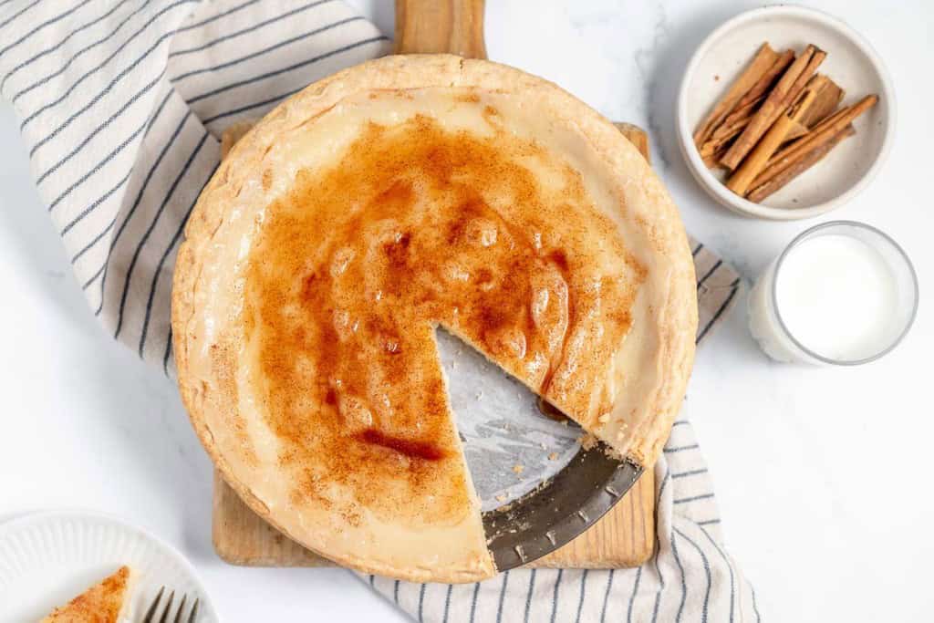 A cream pie with a slice removed, served on a wooden board with a striped cloth, a bowl of cinnamon sticks, and a glass of milk nearby.