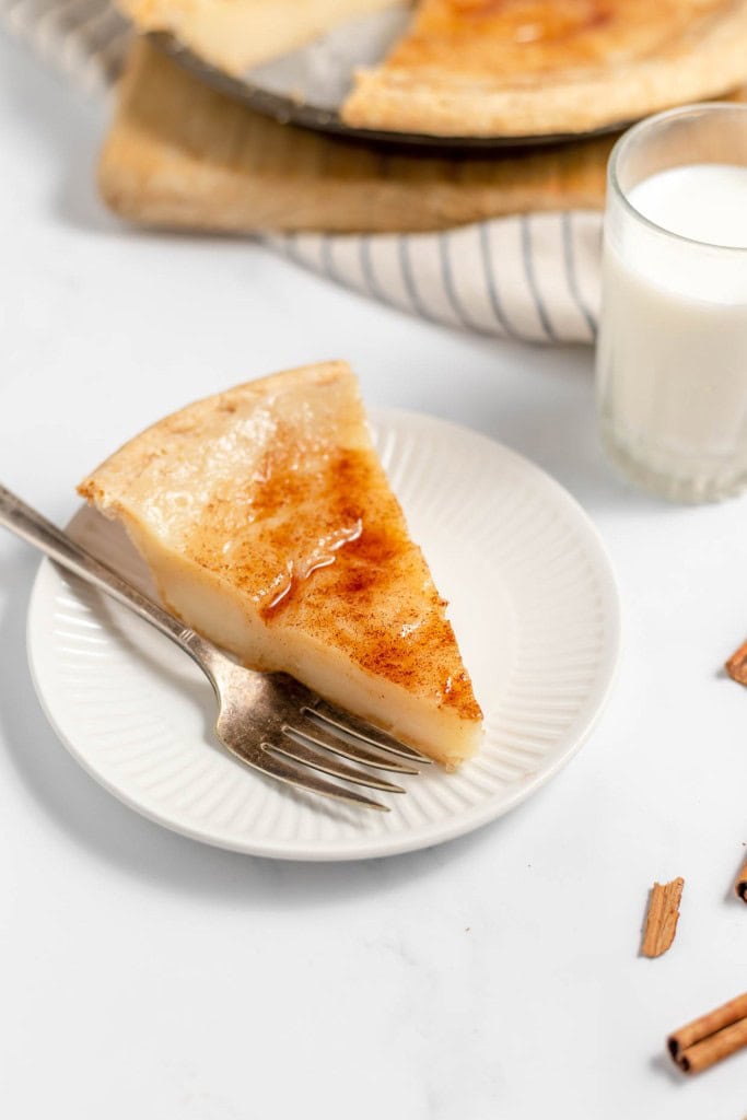 A slice of custard pie on a white plate with a fork, served next to a glass of milk.
