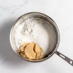 A saucepan containing white flour, white sugar, and light brown sugar, positioned on a white marble surface.