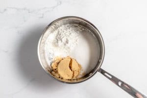 A saucepan containing white flour, white sugar, and light brown sugar, positioned on a white marble surface.