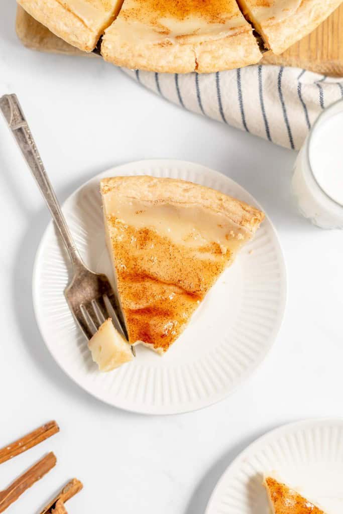 A slice of pie with a fork on a white plate, next to a glass of milk, a whole pie, and some cinnamon sticks on a white surface.