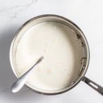 A saucepan filled with milk and a spoon rests inside it. The milk appears to have been recently heated, and there are a few bubbles and possible residue on the surface.